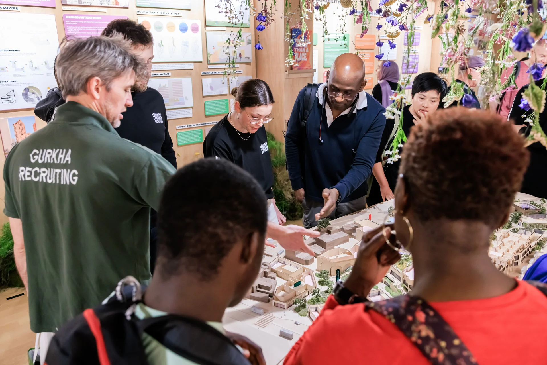 A diverse group examines an architectural model in a well-lit room with informational displays and hanging floral decorations.