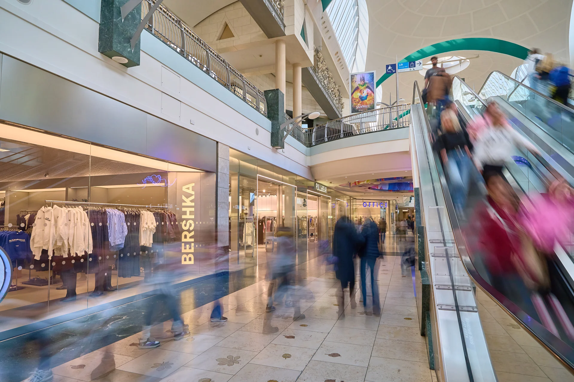 Long exposure interior view of a **Bluewater Shopping Centre** mall corridor, showing the **Bershka** store and shoppers ascending and descending on an escalator with motion blur.