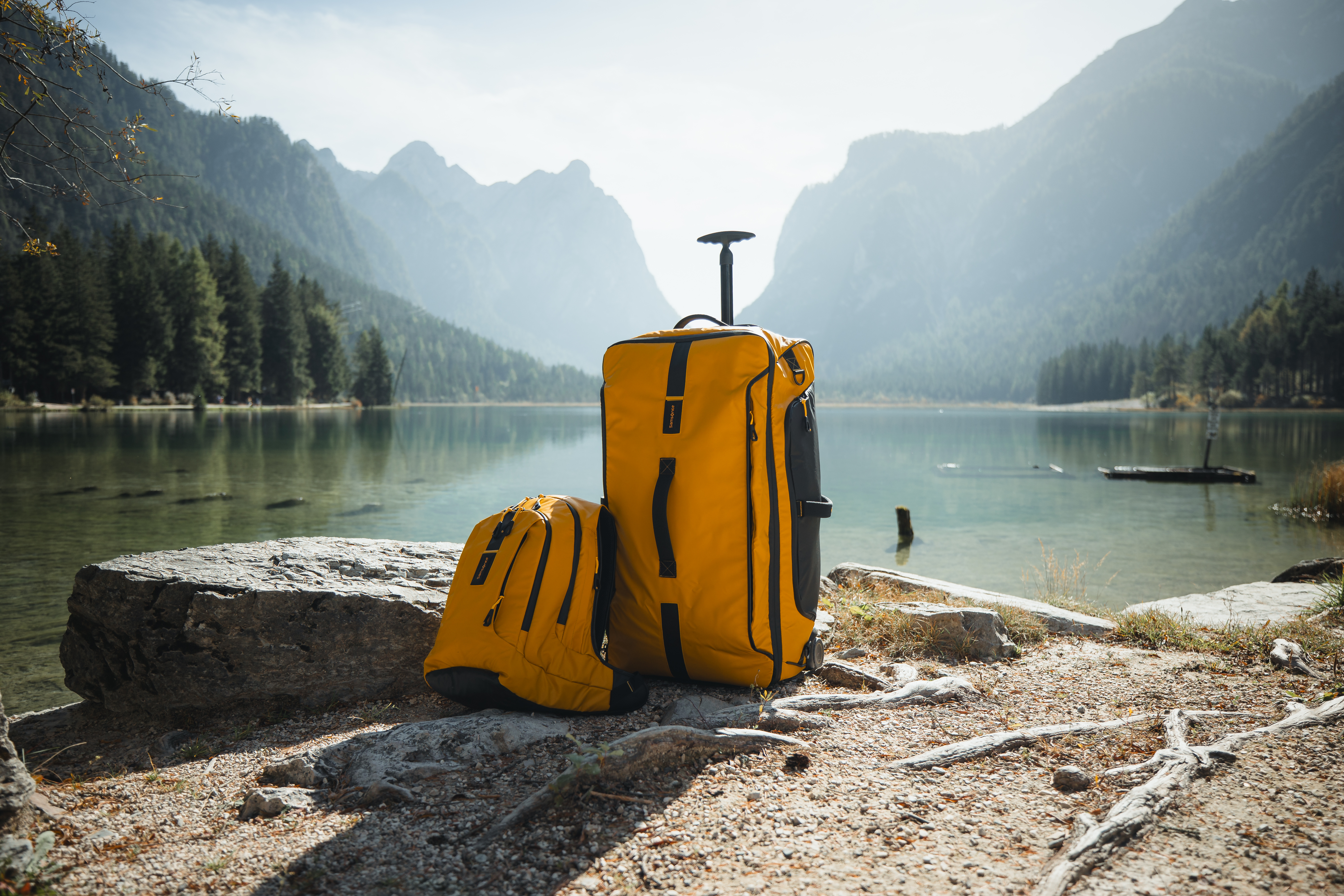 A yellow and black fabric Samsonite suitcase and drawstring bag on a rocky ground in front of a lake and mountains
