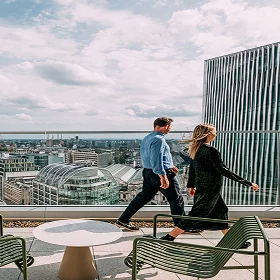 A man and a woman walk across a modern **rooftop terrace** with lounge chairs, overlooking the panoramic skyline of a city, featuring contemporary architecture.