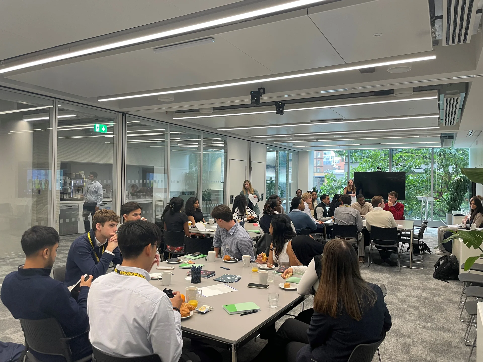 A group of **young professionals** or students participating in a large **business workshop** or corporate training session in a modern, glass-walled conference room, with attendees seated around tables and taking notes.
