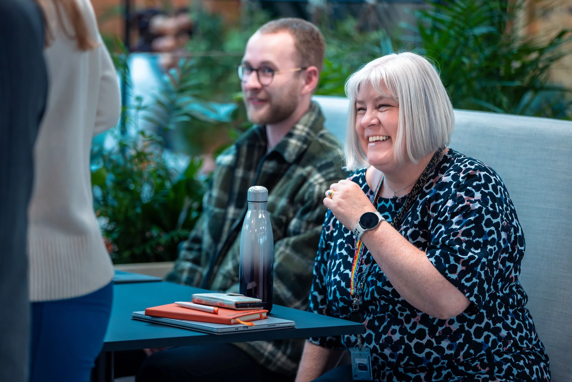 Two people seated at a table with notebooks and a water bottle, smiling during a meeting in a green, indoor space.