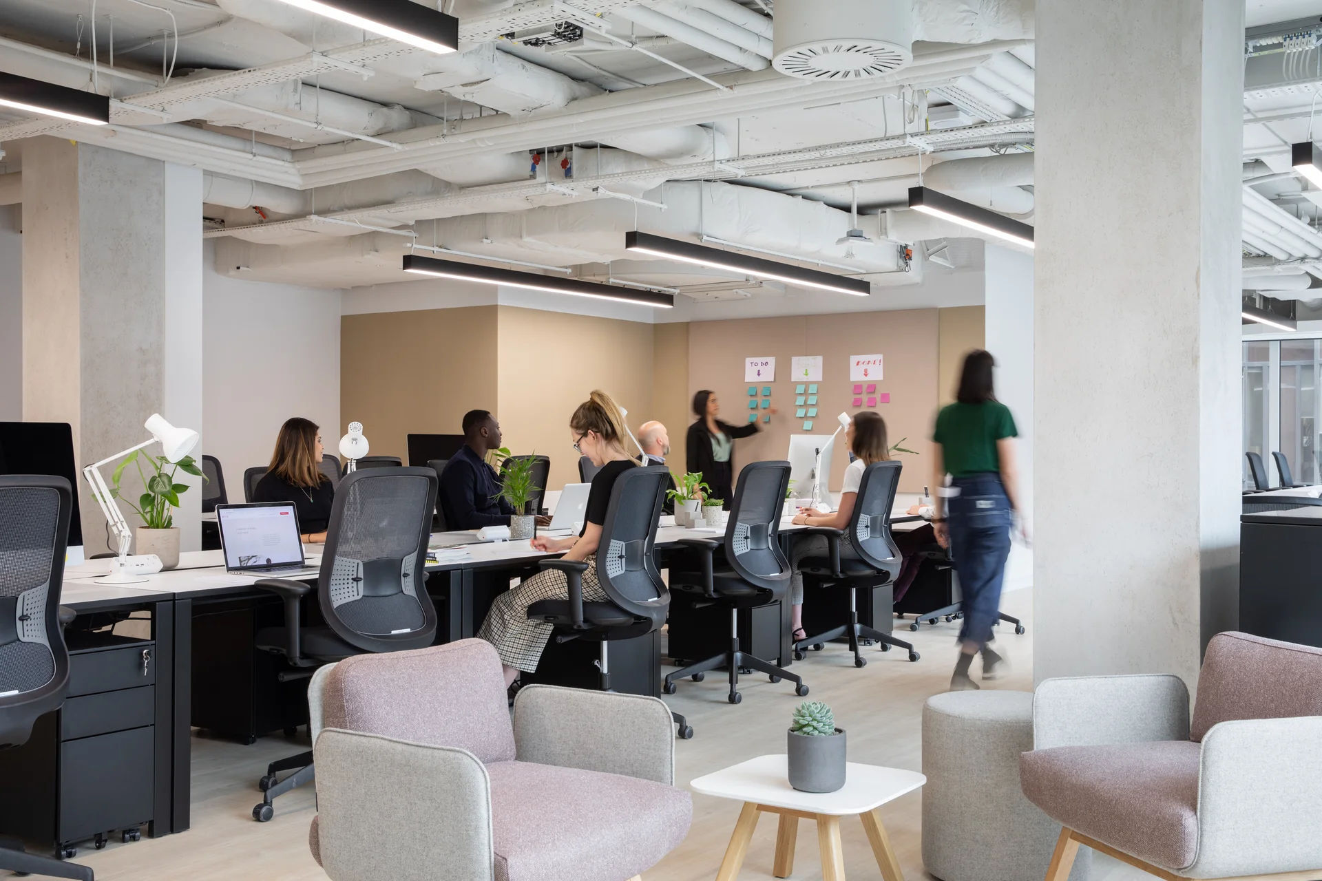 Modern office space with open desks, workers seated at computers, and a casual seating area with chairs and a small table.