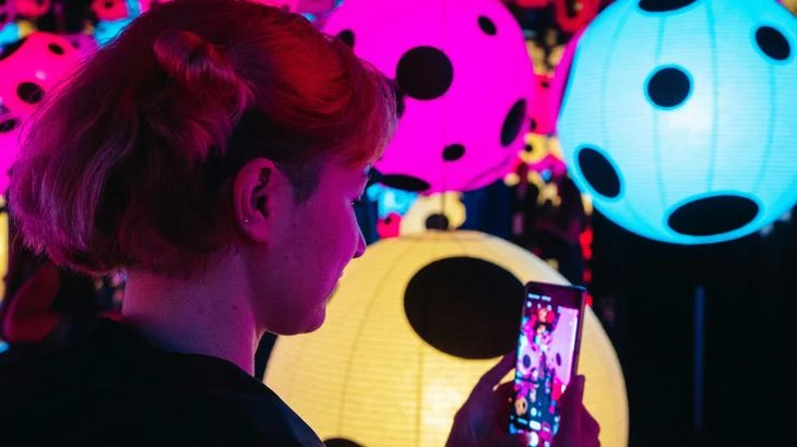 Person with red hair looking at a smartphone with colorful, spotted lanterns in the background.