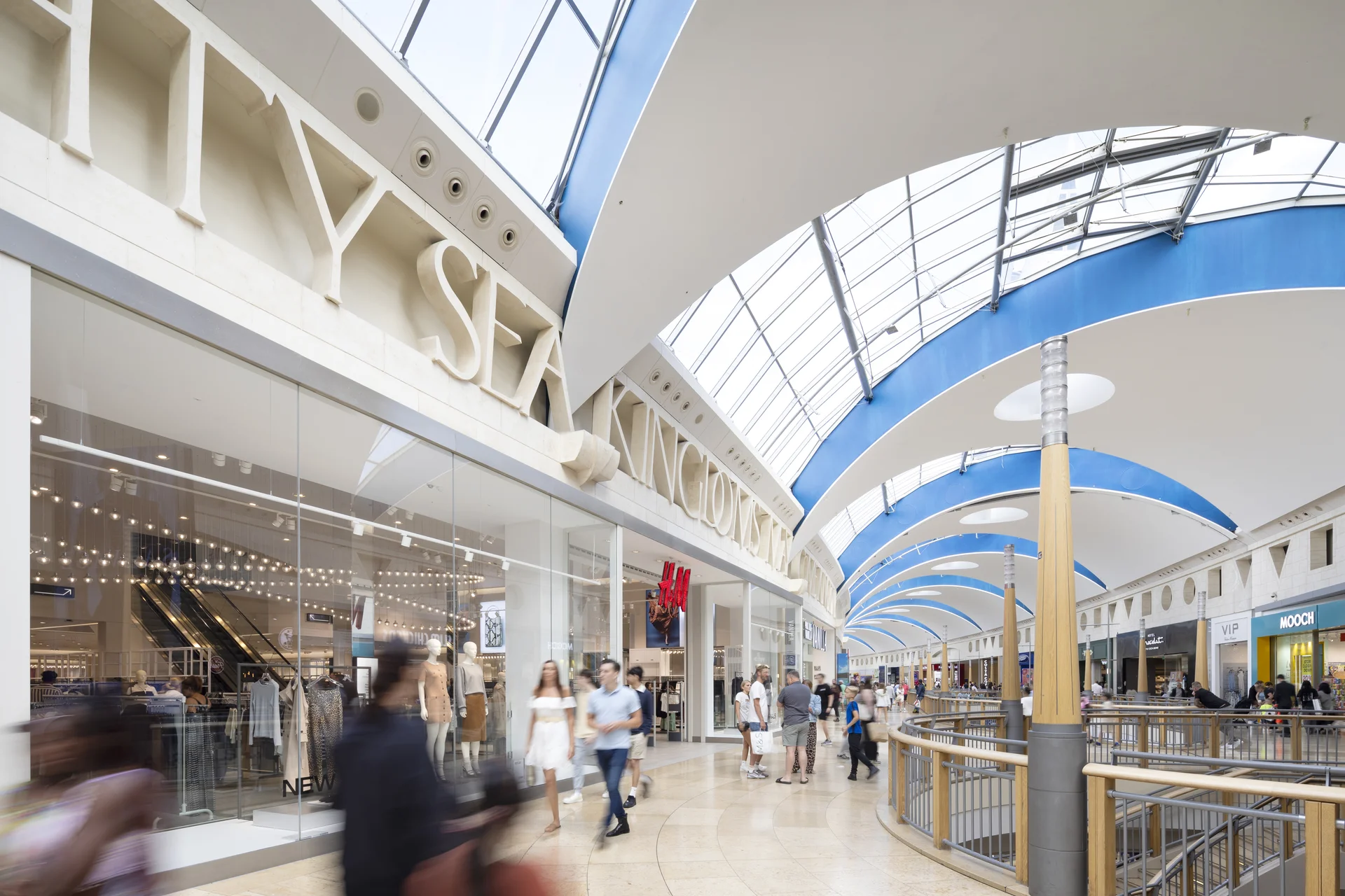 Interior view of the **Bluewater Shopping Centre** mall, featuring high curved ceilings, skylights, and shoppers walking past large storefronts like H&M, with a sense of movement blur.