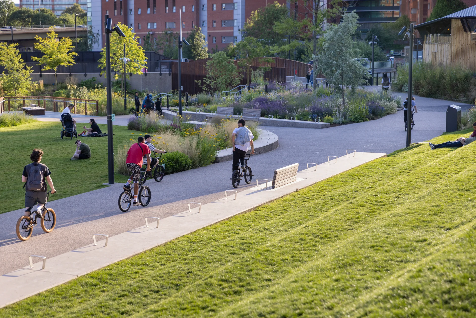 People biking and relaxing in a green urban park with modern architecture and lush landscaping in the background.