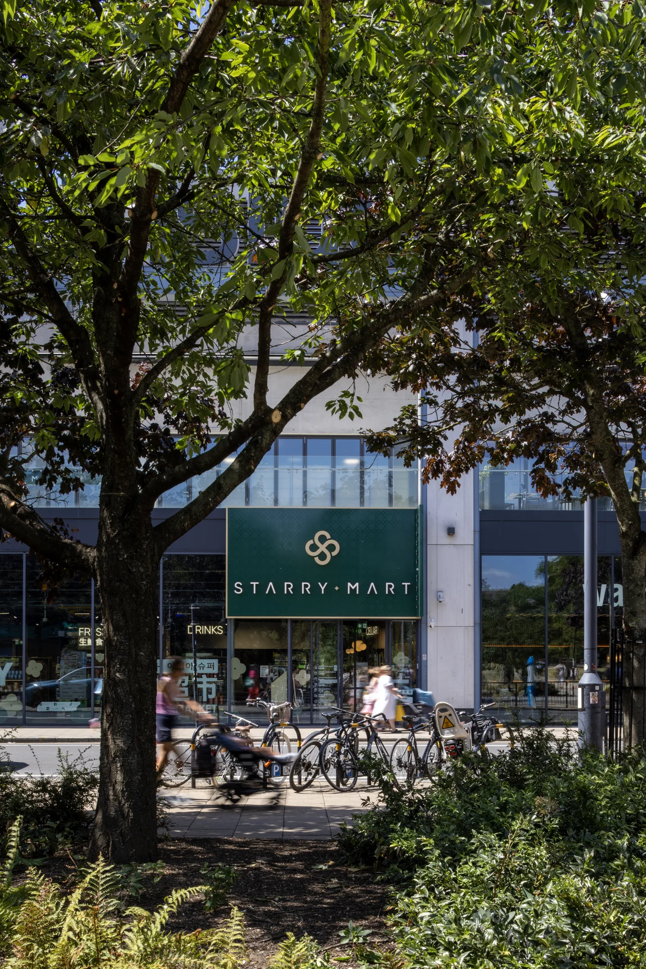 Exterior view of a **Starry Mart** storefront at **Southside** through the foliage of large trees, with a row of parked bicycles and motion-blurred pedestrians and cyclists.