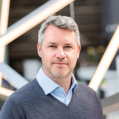 Bruce Findlay in gray sweater and blue shirt against geometric background.