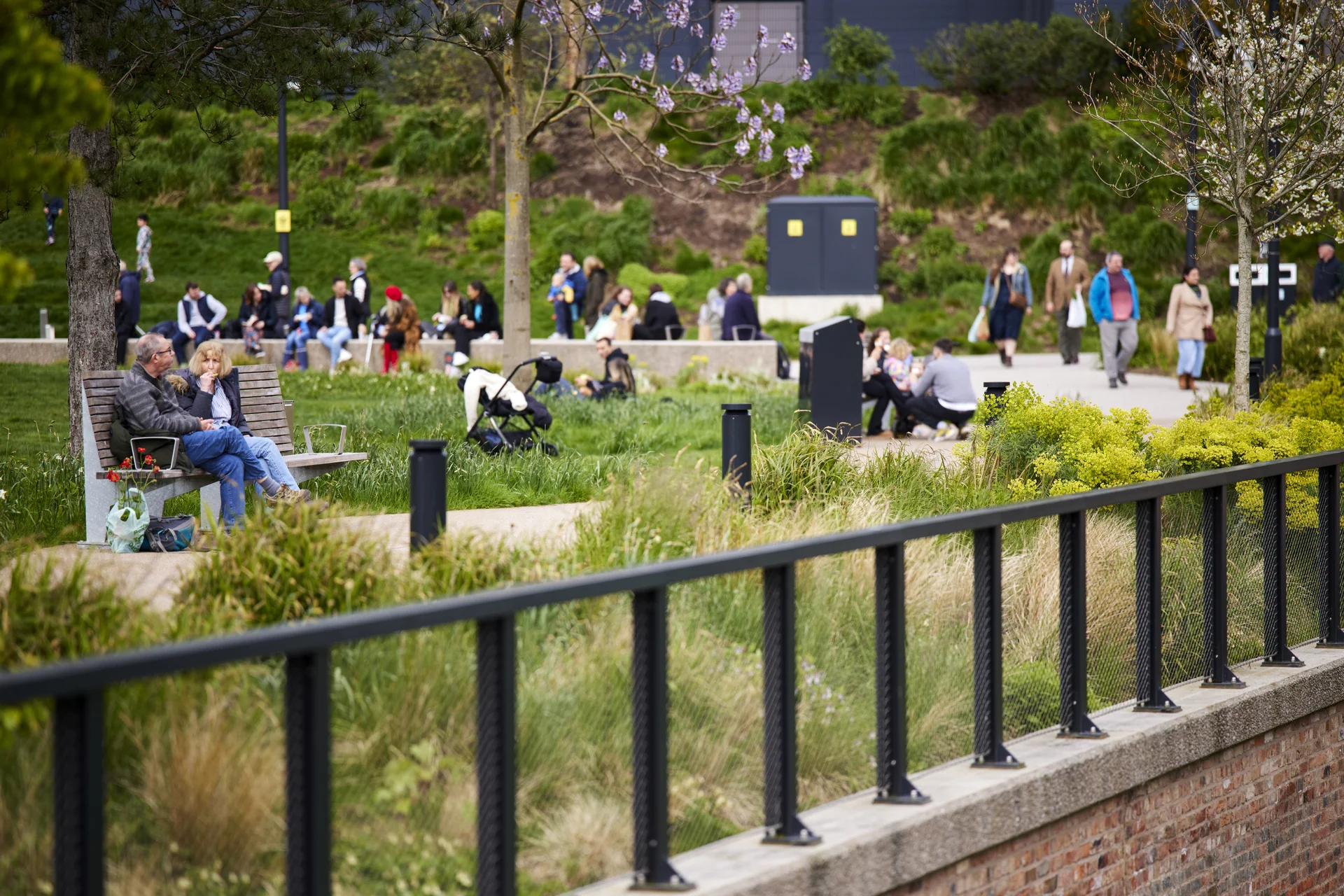 A view of a busy urban green space on a sunny day, showing people sitting on benches and grassy slopes, walking, and relaxing in a modern public park setting.