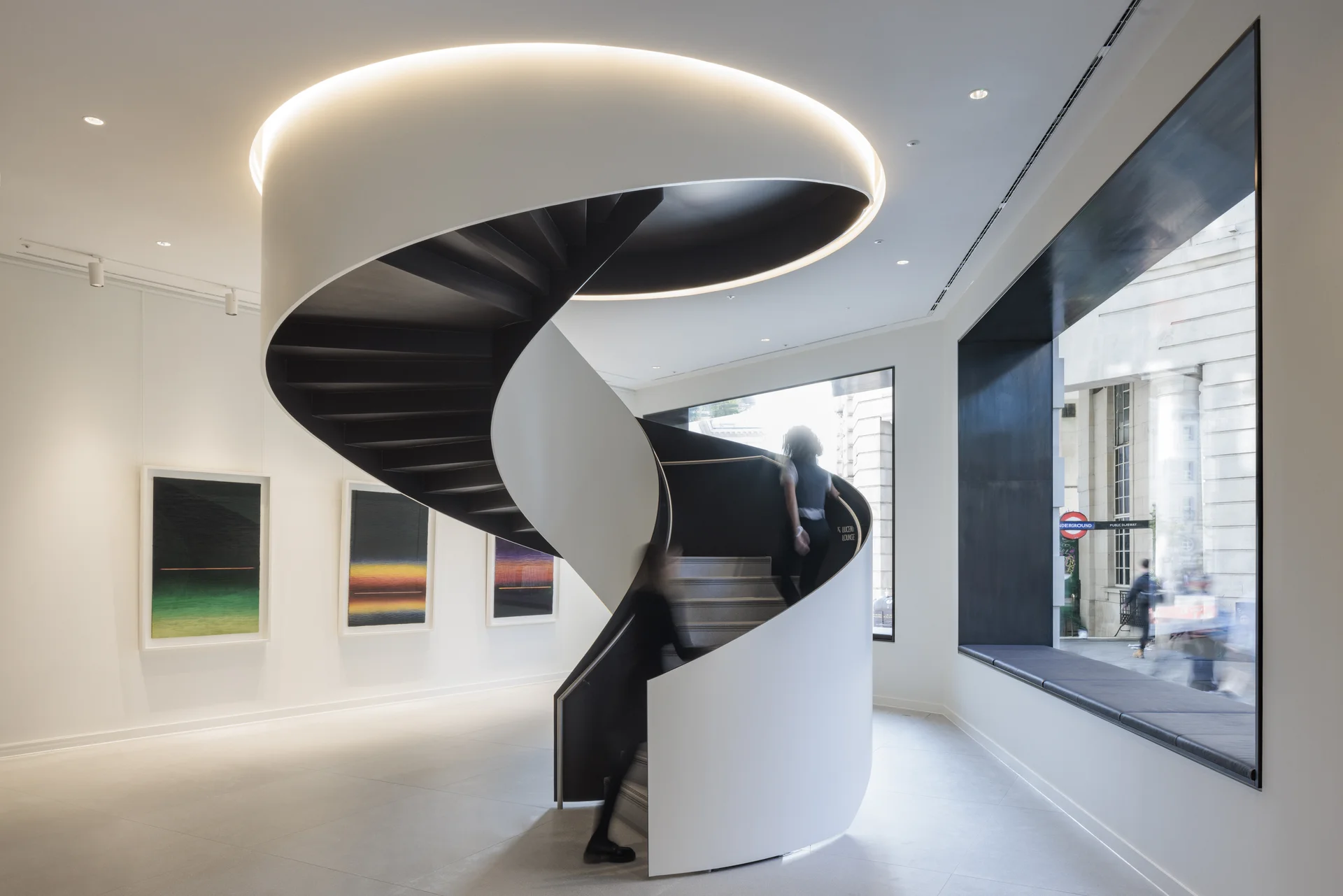 A modern spiral staircase with black steps and white railing in a bright gallery space with artwork and large windows.