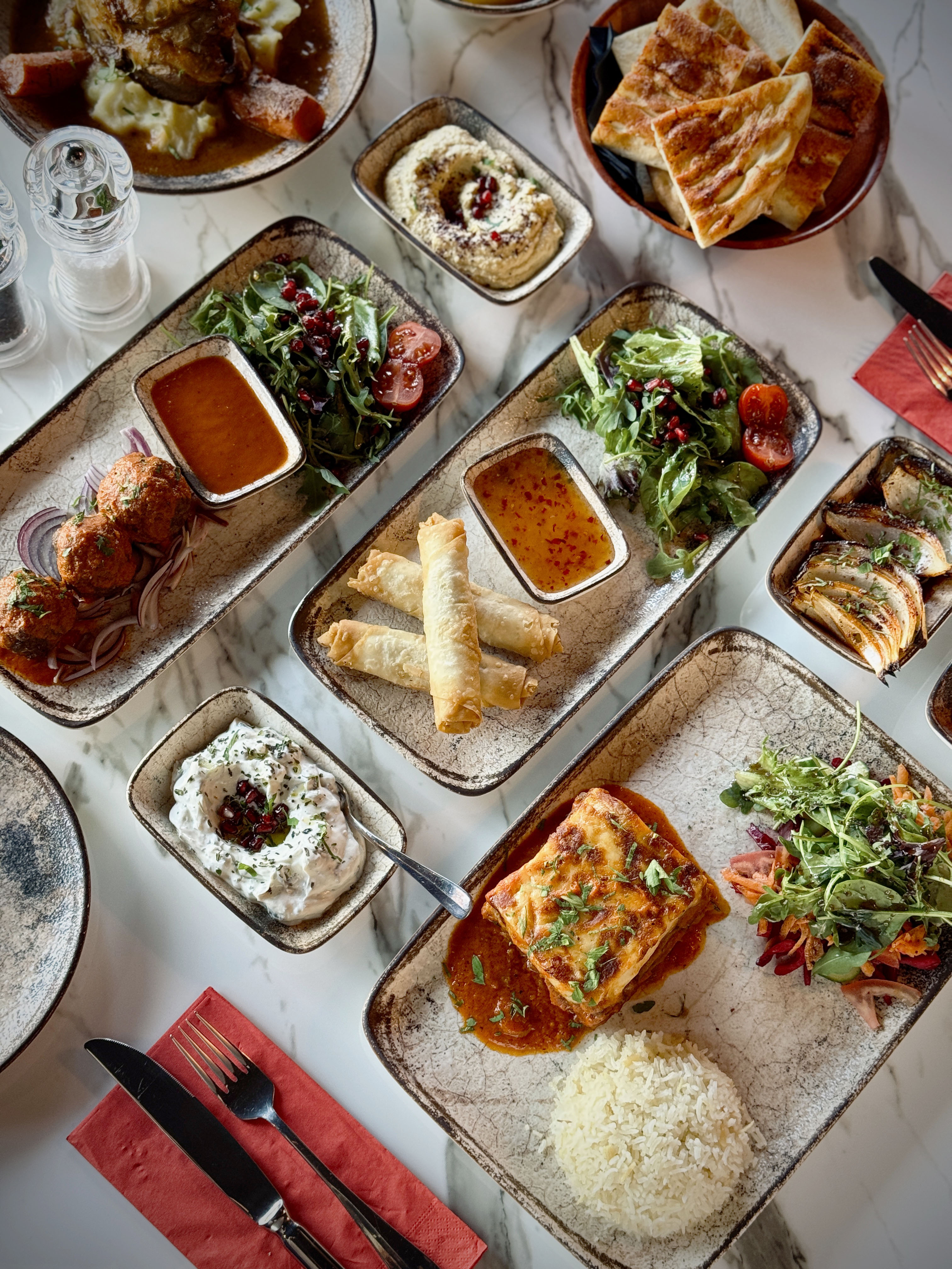 An array of Mediterranean dishes on a marble table, including hummus, dolmas, borek, salad, and bread. Presentation is colorful and appetizing.