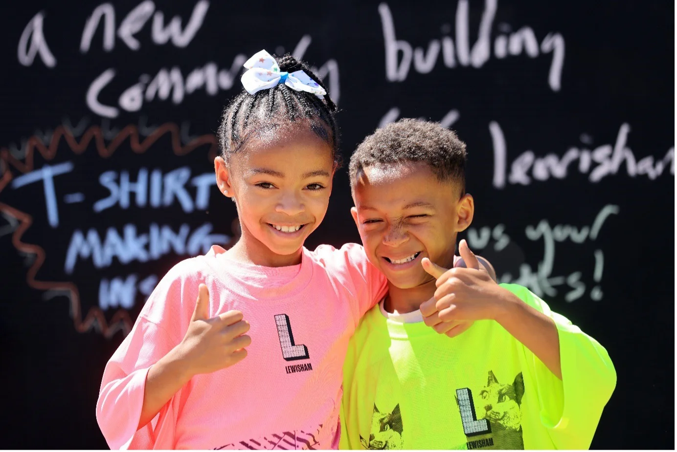 Two children giving thumbs up in front of a chalkboard with community event text, wearing bright shirts.