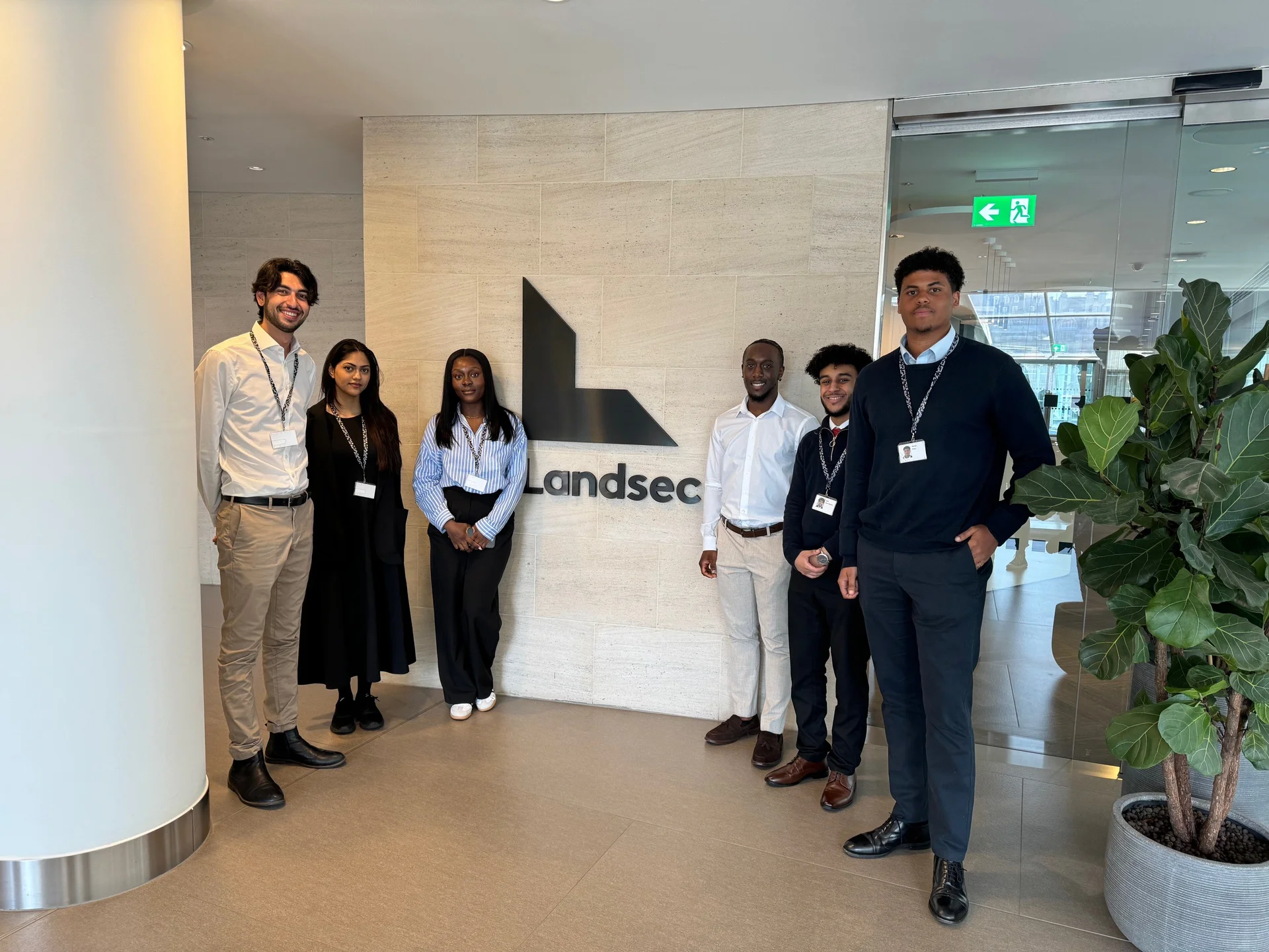 Six individuals stand in a modern office lobby, posing in front of a 'Landsec' logo on a light stone wall, with glass doors and a plant nearby.