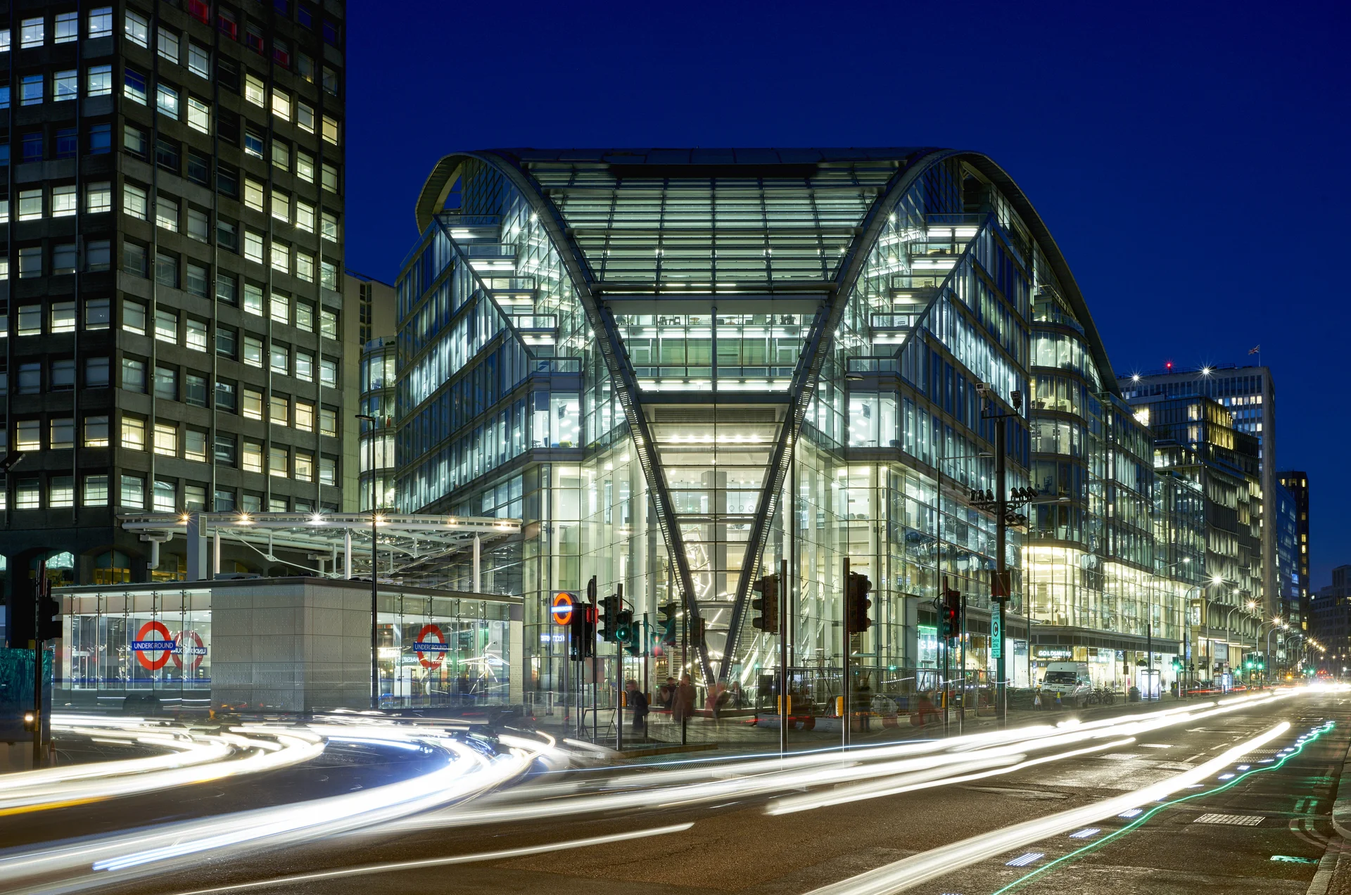 Modern glass building with arched roof at dusk, surrounded by urban structures and blurred traffic lights.