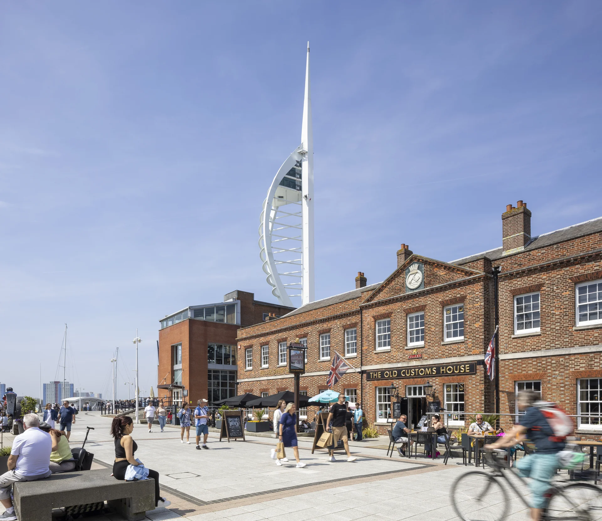 View of **Gunwharf Quays** in Portsmouth featuring the historic **The Old Customs House** brick building in the foreground and the modern, iconic **Spinnaker Tower** dominating the background skyline.
