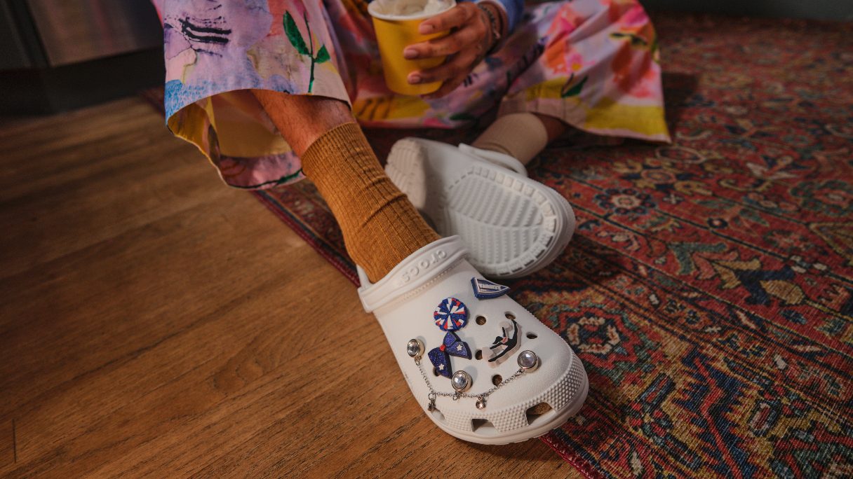 A person wearing colourful trousers with white, decorated Crocs sits on a boho carpet.